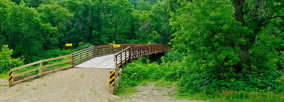 Entrance to walking, biking bridge in a park surrounded by green, leafy trees