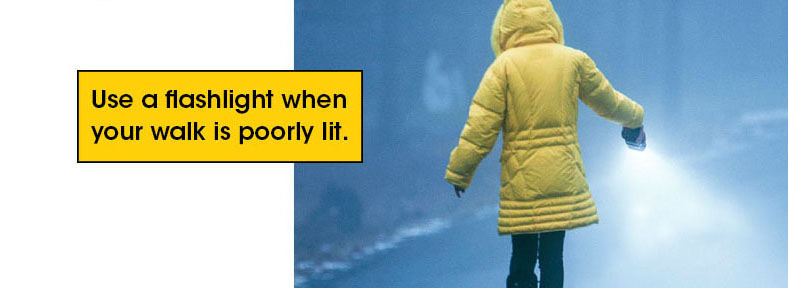 Woman in yellow winter coat walks along dark path using a flashlight to light the way. Text; Use a flashlight when your walk is poorly lit.