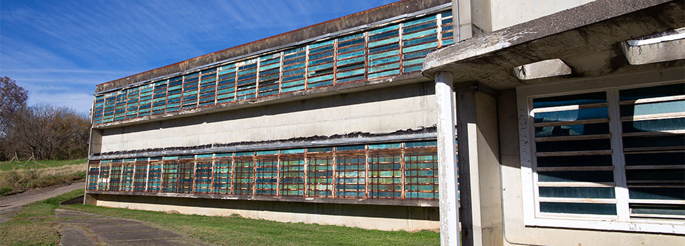 Abandoned commercial building. Concrete falling apart, windows rusty and sidewalk overgrown with grass.