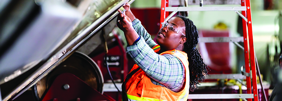 A mature African-American woman working in a metal fabrication shop. She is wearing a safety vest and protective goggles, using a tape measure to measure part of a large metal object.