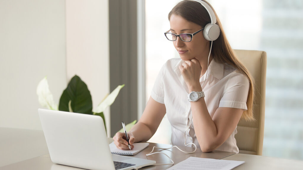 Focused attentive woman in headphones sits at desk with laptop, looks at screen, makes notes, learns foreign language in internet, online study course, self-education on web, consults client by video
